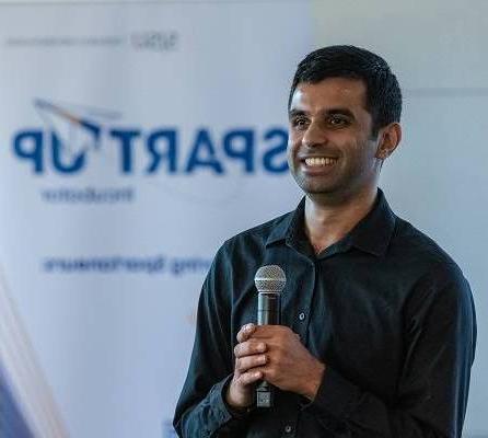 A dark-haired man holds a microphone and smiles, with a white pop-up banner that says "SpartUp Incubator" behind him.